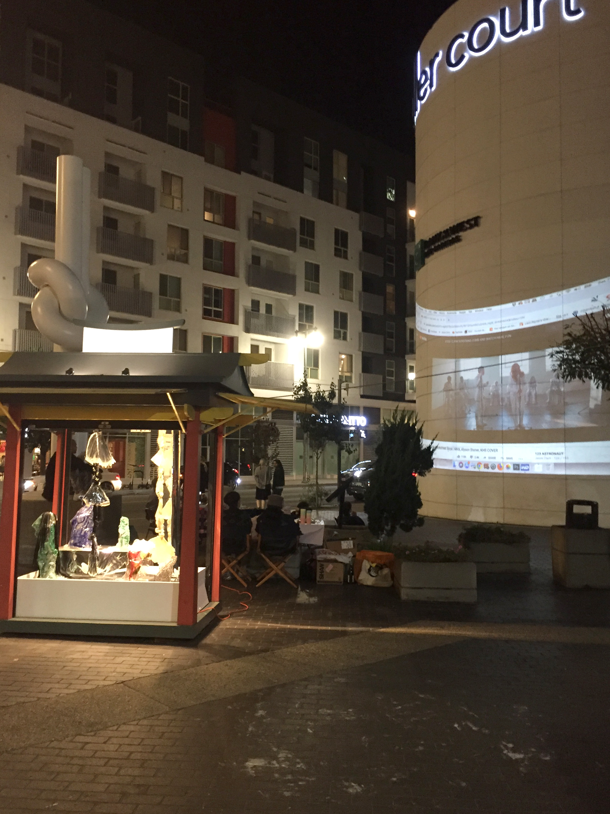 A corner in Little Tokyo, Los Angeles with a projection on a bank building behind a cell phone kiosk full of art